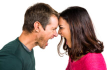 A man and a woman are facing each other with their foreheads touching, both yelling aggressively. The man is wearing a dark green shirt, and the woman is wearing a bright pink sweater. The background is plain white.