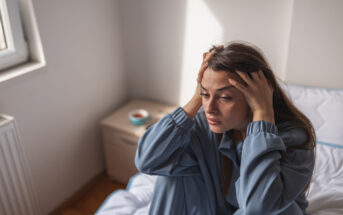 A woman in blue pajamas sits on a bed with her hands on her head, looking distressed. The room has white walls, a window, and a small bedside table with a bowl and a cup. Natural light illuminates the room.