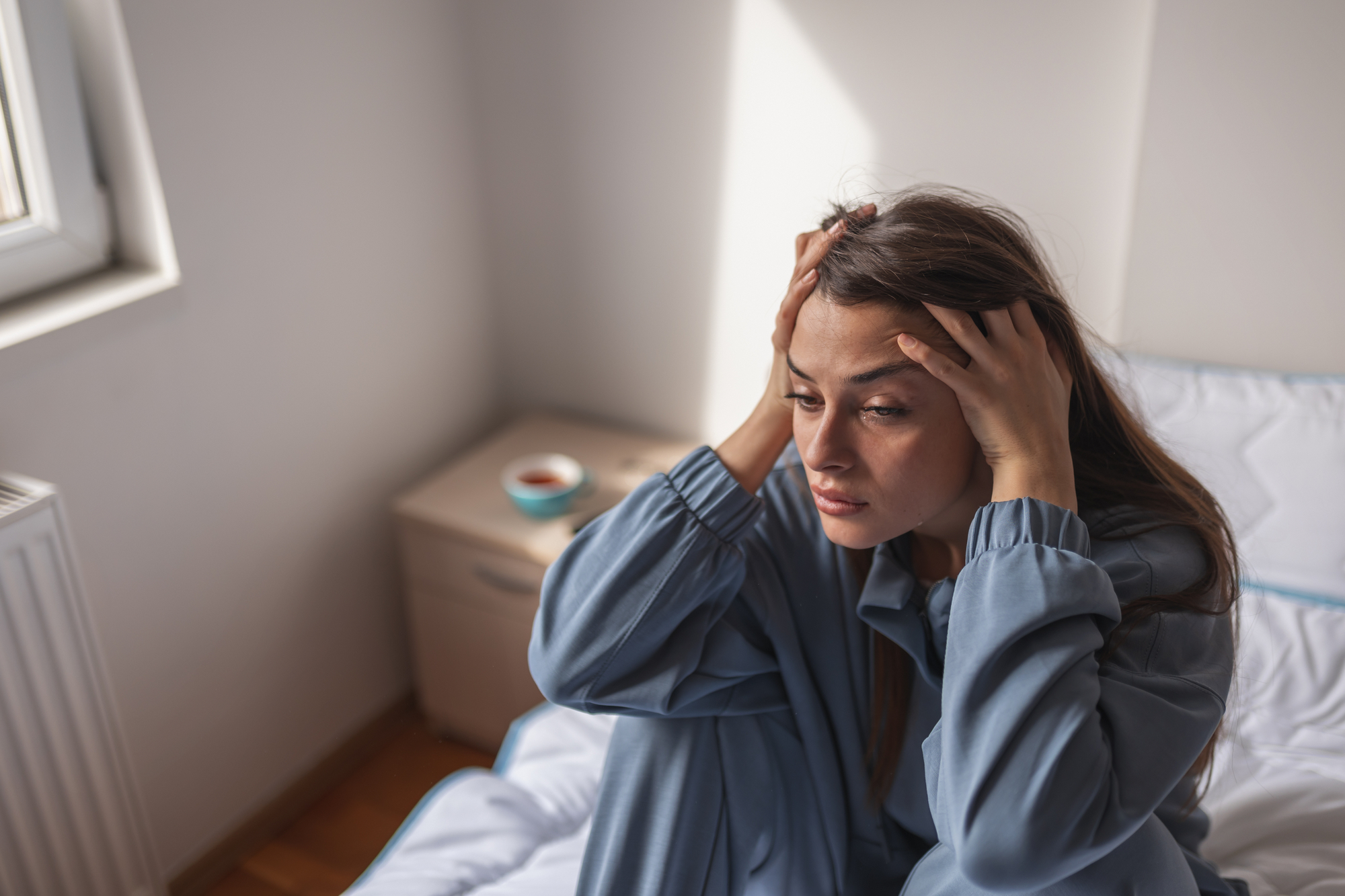 A woman in blue pajamas sits on a bed with her hands on her head, looking distressed. The room has white walls, a window, and a small bedside table with a bowl and a cup. Natural light illuminates the room.