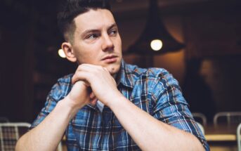 A young man with short dark hair, wearing a blue plaid shirt, sits at a table with his hands clasped in front of him. He is looking to his right with a thoughtful expression. The background is softly lit with warm lighting and out-of-focus elements.