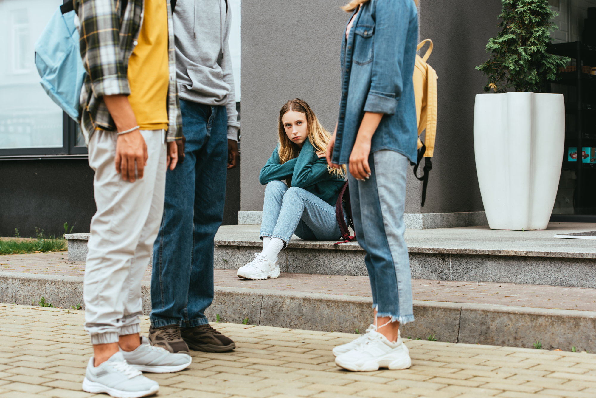 A girl sits alone on outdoor steps, looking distressed, while three standing people face each other nearby. The seated girl wears a green jacket and jeans. The standing individuals wear casual clothes and backpacks, their faces are not shown.