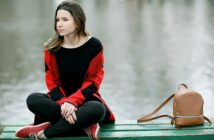 A person with long hair sits on a green wooden bench near the water, wearing a red and black sweater, black pants, and red shoes. They have a contemplative expression. A small brown backpack is placed beside them on the bench.