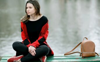 A person with long hair sits on a green wooden bench near the water, wearing a red and black sweater, black pants, and red shoes. They have a contemplative expression. A small brown backpack is placed beside them on the bench.