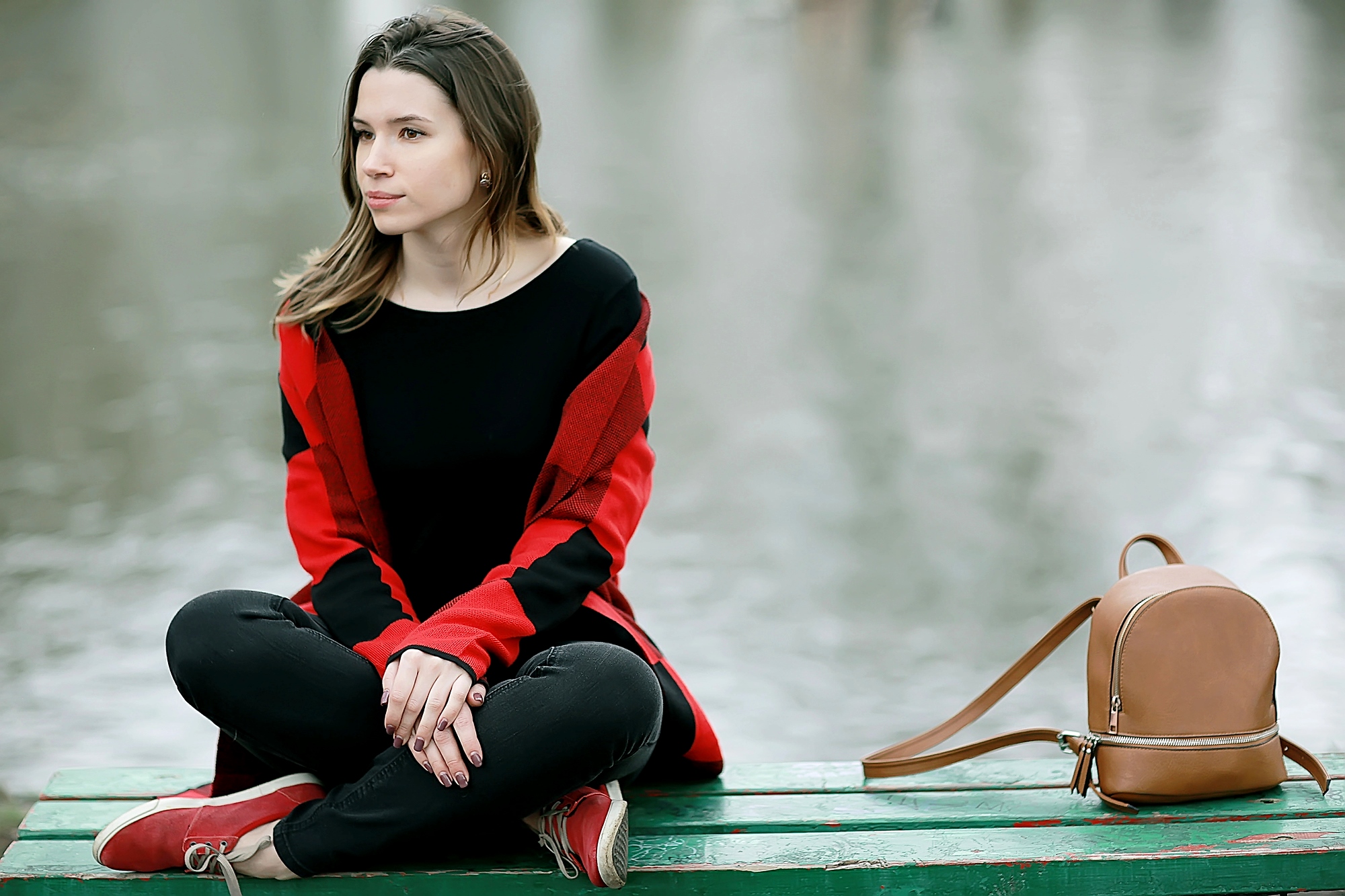 A person with long hair sits on a green wooden bench near the water, wearing a red and black sweater, black pants, and red shoes. They have a contemplative expression. A small brown backpack is placed beside them on the bench.