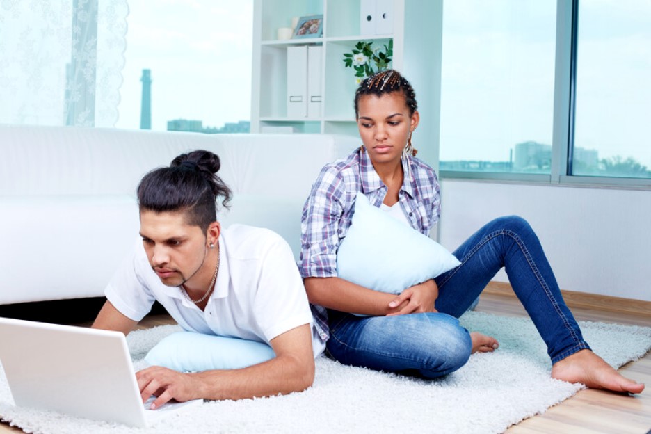 A man lying on a carpeted floor types on a laptop while a woman sits next to him, leaning against a couch and holding a pillow. They appear to be in a bright, modern living room with large windows in the background.