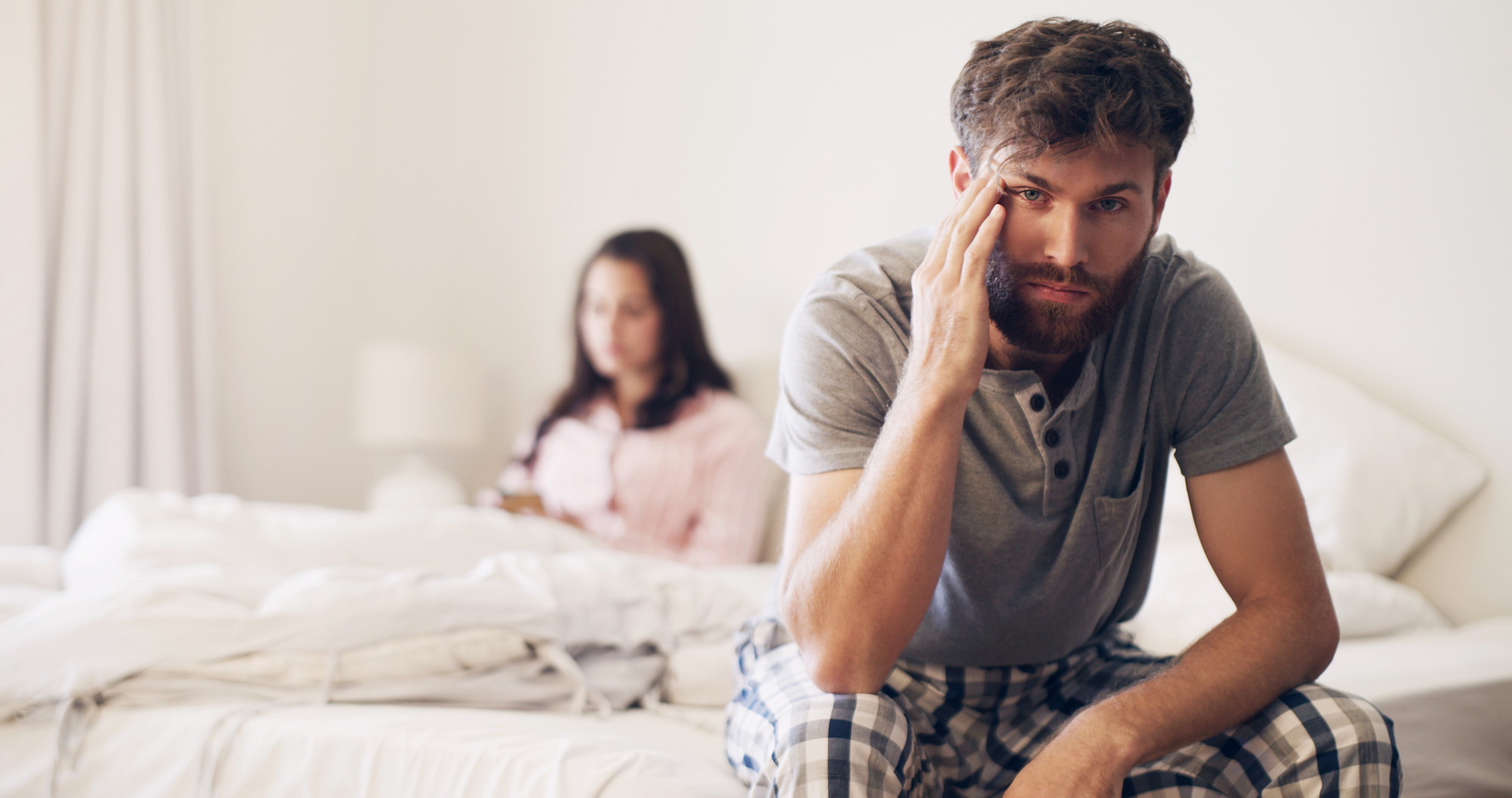 A bearded man in a gray shirt and plaid pajama bottoms sits on the edge of a bed, looking distressed with his hand on his temple. A woman in the background, wearing a pink shirt, sits on the bed facing away, creating a tense atmosphere.