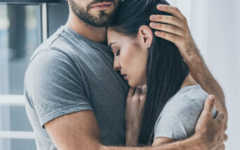 A close-up of a couple hugging each other. The man, with a beard, is wearing a gray t-shirt, and he has one hand gently placed on the woman's head. The woman, also in a gray shirt, has her head resting on the man's chest with a serene expression.