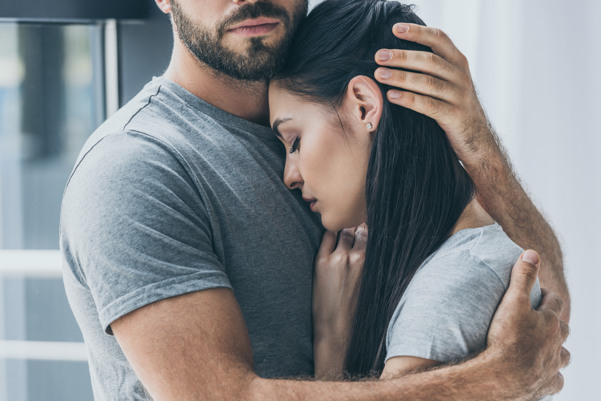 A close-up of a couple hugging each other. The man, with a beard, is wearing a gray t-shirt, and he has one hand gently placed on the woman's head. The woman, also in a gray shirt, has her head resting on the man's chest with a serene expression.