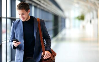 A man with short brown hair, wearing a blue blazer and black shirt, smiles while looking at his smartphone. He has a brown leather shoulder bag and is walking in a spacious, modern corridor with large windows and natural light.