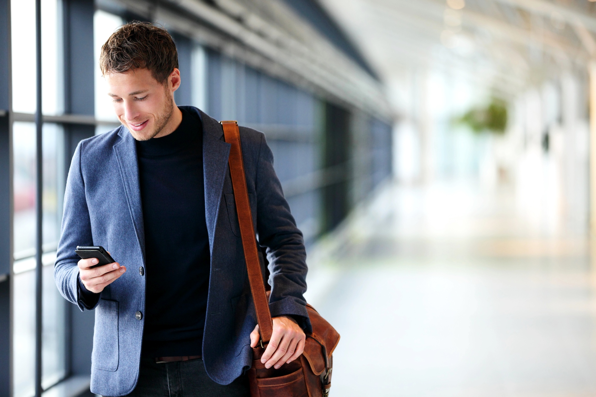 A man with short brown hair, wearing a blue blazer and black shirt, smiles while looking at his smartphone. He has a brown leather shoulder bag and is walking in a spacious, modern corridor with large windows and natural light.