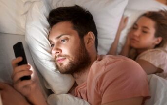 A man with a beard is lying in bed, propped up by pillows, and is looking at a smartphone he is holding. Beside him, a woman is sleeping peacefully, also snuggled in the bed with white sheets and pillows. Both are wearing casual clothes.