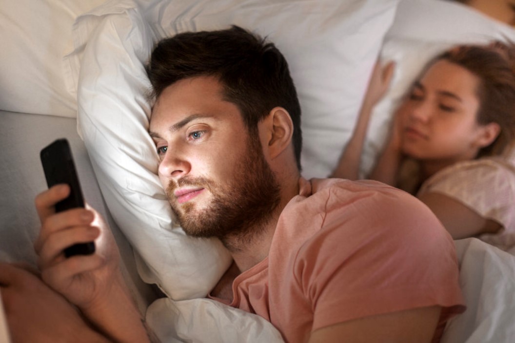 A man with a beard is lying in bed, propped up by pillows, and is looking at a smartphone he is holding. Beside him, a woman is sleeping peacefully, also snuggled in the bed with white sheets and pillows. Both are wearing casual clothes.