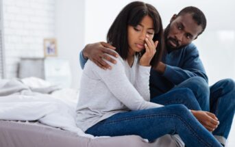 A man sits on a bed, holding a woman who appears to be upset. The woman, wearing a light gray sweater and jeans, has her hand to her face with eyes closed, while the man, wearing a dark blue sweater and jeans, gently places his arm around her shoulder.