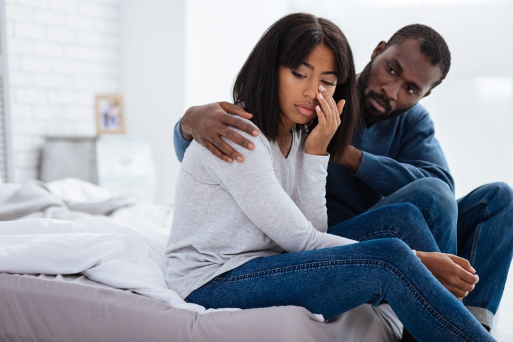 A man sits on a bed, holding a woman who appears to be upset. The woman, wearing a light gray sweater and jeans, has her hand to her face with eyes closed, while the man, wearing a dark blue sweater and jeans, gently places his arm around her shoulder.