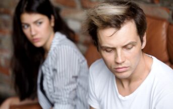 A man with light brown hair, wearing a white t-shirt, looks down with a serious expression. Behind him, a woman with long dark hair and a striped shirt looks at him with a concerned expression. They are indoors with a brick wall in the background.