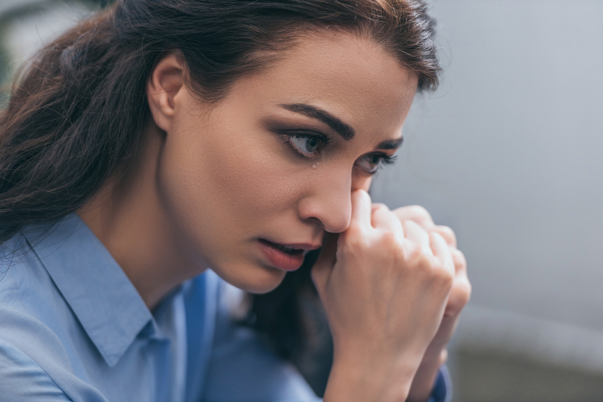 A close-up of a woman with long dark hair and a blue shirt. She looks pensive and concerned, resting her chin on her clasped hands while gazing into the distance, deep in thought. The background is blurred, emphasizing her expression and emotions.