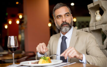 A distinguished man with a beard and mustache, wearing a beige suit and a navy polka-dot tie, is seated at a restaurant table. He is holding a fork and knife, about to eat from a plated meal. A glass of red wine is on the table beside him.