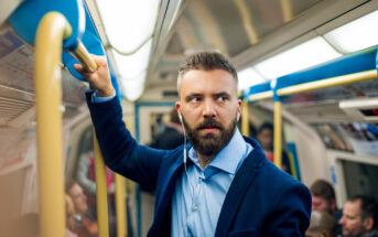 A man with a beard, wearing a blue shirt and navy blazer, stands in a crowded subway car, holding onto a yellow handrail. He has earphones in and appears to be looking out of the frame. Other passengers are seated and standing around him.