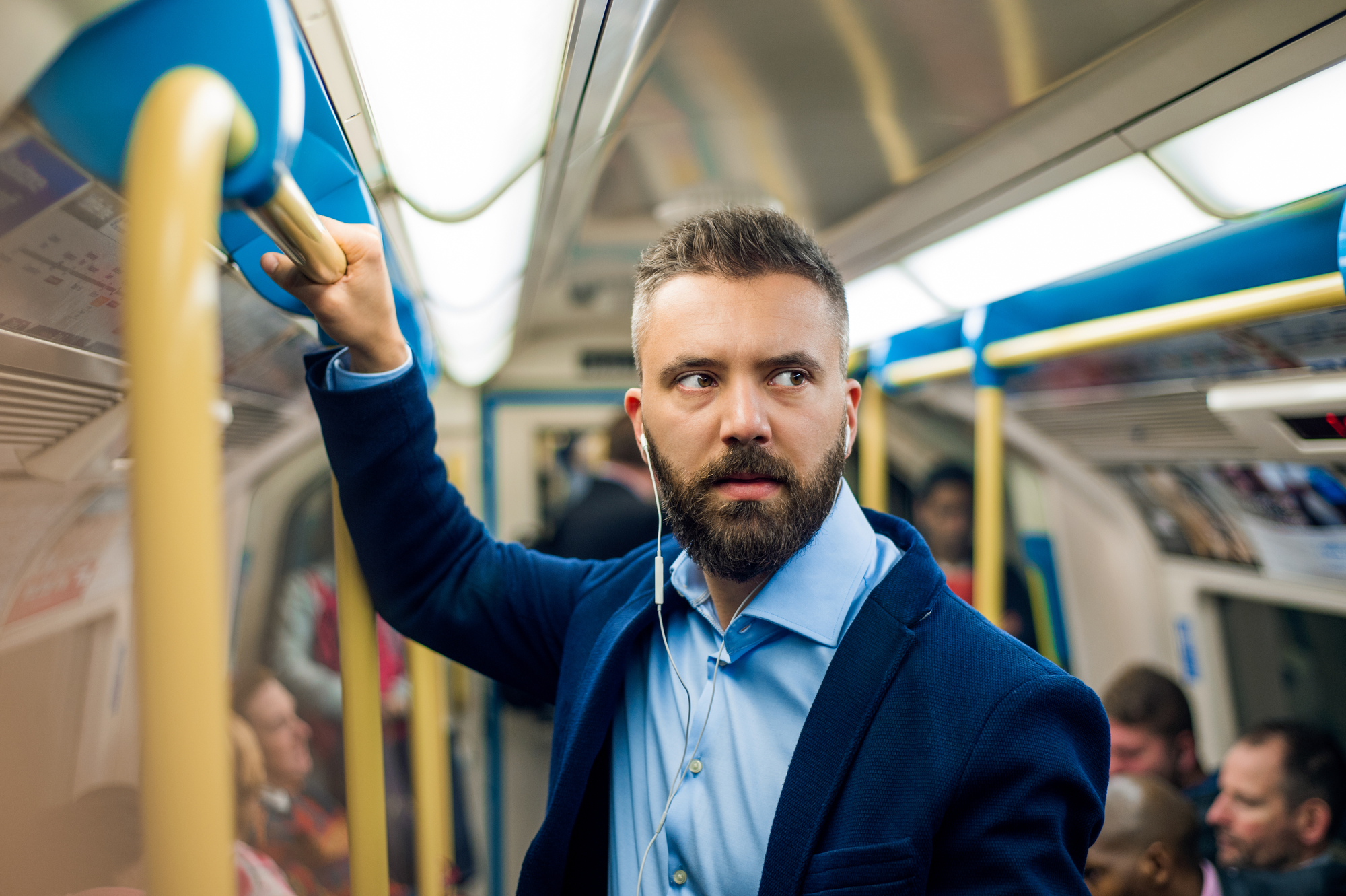 A man with a beard, wearing a blue shirt and navy blazer, stands in a crowded subway car, holding onto a yellow handrail. He has earphones in and appears to be looking out of the frame. Other passengers are seated and standing around him.