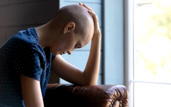A person with a bald head, wearing a dark blue shirt with white polka dots, is sitting inside and leaning forward with their hand resting on their head. They appear to be deep in thought or contemplation near a window with natural light coming in.