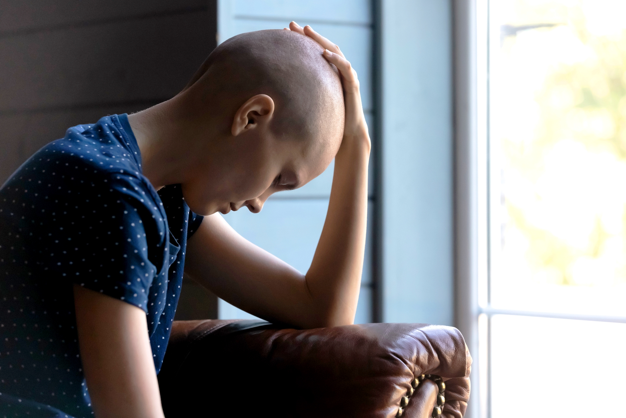 A person with a bald head, wearing a dark blue shirt with white polka dots, is sitting inside and leaning forward with their hand resting on their head. They appear to be deep in thought or contemplation near a window with natural light coming in.