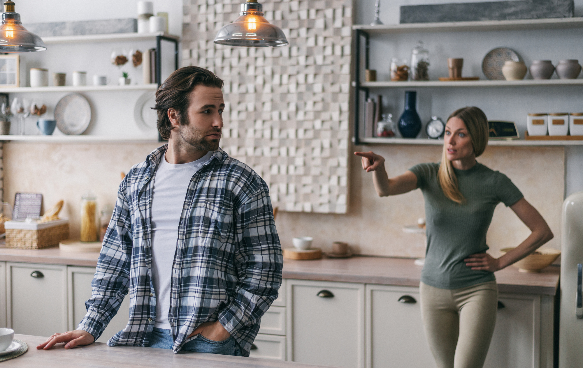 A man leaning against a kitchen counter, wearing a plaid shirt, looks away with a contemplative expression. A woman standing behind him is pointing and appears to be speaking to him sternly. The kitchen has modern decor with open shelves and various dishes.