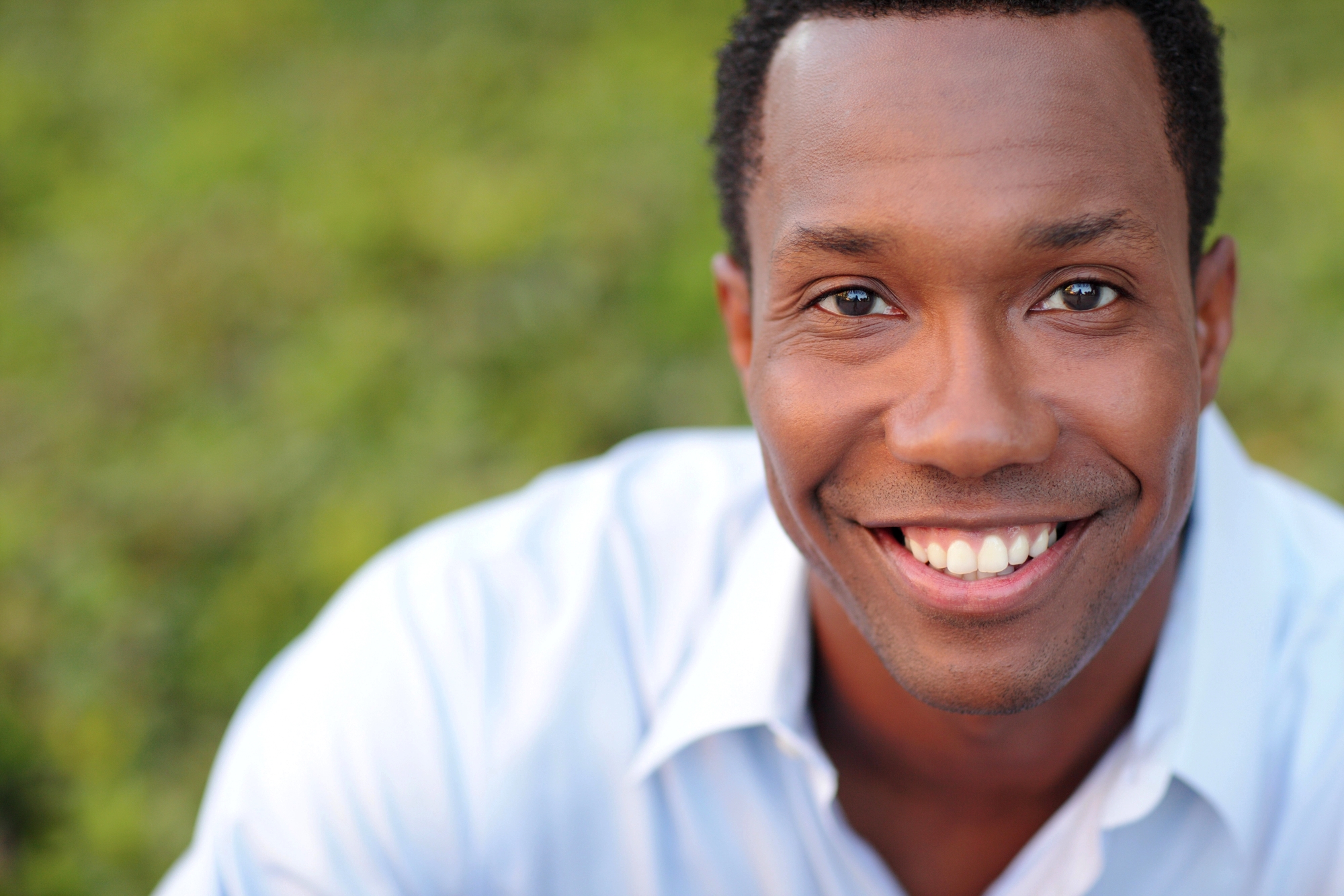 A man with short curly hair is smiling at the camera. He is wearing a light blue collared shirt. The background is blurred greenery.