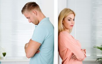 A man and a woman stand back-to-back with a wall separating them. Both have their arms crossed and appear upset. The man wears a light blue shirt, and the woman wears a peach-colored blouse. The background shows white blinds partially covering a window.