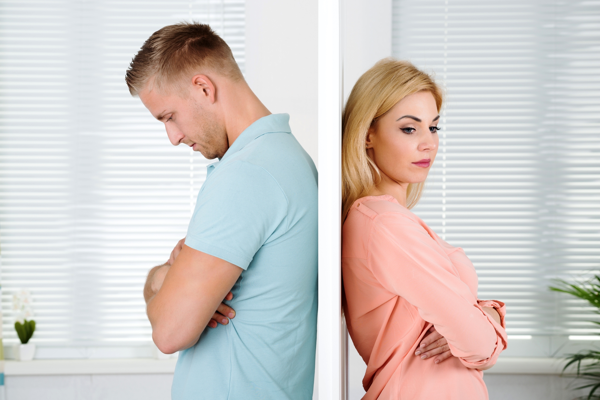 A man and a woman stand back-to-back with a wall separating them. Both have their arms crossed and appear upset. The man wears a light blue shirt, and the woman wears a peach-colored blouse. The background shows white blinds partially covering a window.