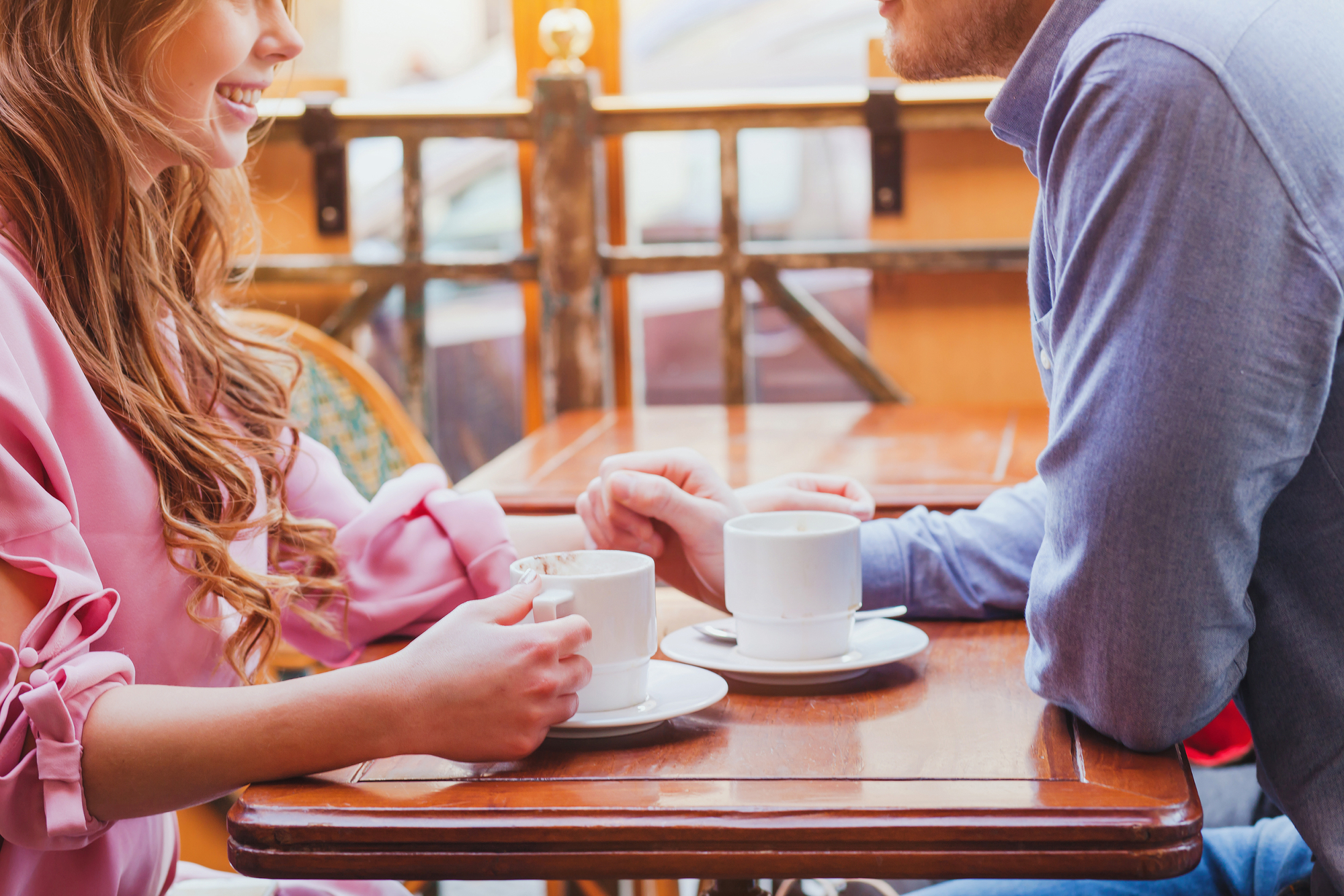 A woman and man are sitting at a table in a cafe, each holding a cup of coffee. The woman is wearing a pink blouse and smiling, and the man is in a blue shirt. They are engaged in conversation, and the background shows an outdoor seating area with blurred details.