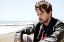 A man with wavy brown hair and a beard is sitting on a beach, wearing a leather jacket. He appears deep in thought as he looks slightly downward. The ocean and a clear blue sky are in the background.
