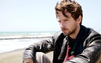 A man with wavy brown hair and a beard is sitting on a beach, wearing a leather jacket. He appears deep in thought as he looks slightly downward. The ocean and a clear blue sky are in the background.