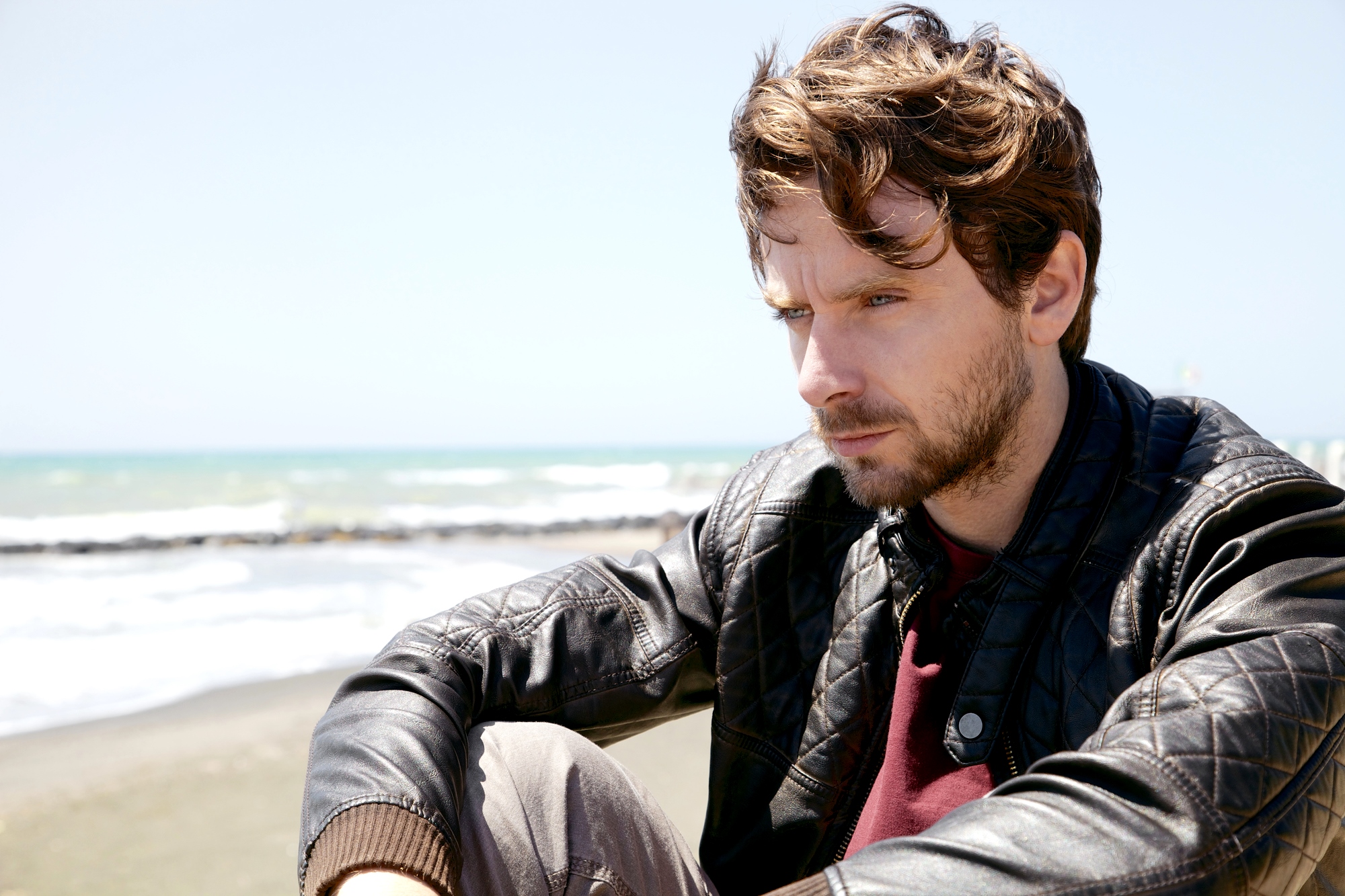 A man with wavy brown hair and a beard is sitting on a beach, wearing a leather jacket. He appears deep in thought as he looks slightly downward. The ocean and a clear blue sky are in the background.