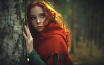 A woman with long, curly red hair leans against a tree in a forest. She is wrapped in a bright red scarf and gazes sideways with a pensive expression. Sunlight filters through the trees, casting a warm glow on the scene.