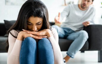 A woman sits on the floor with her head resting on her crossed arms, looking downcast. A man in the background, sitting on a couch, appears upset and is gesturing towards her. The scene suggests a conflict or argument between the two.