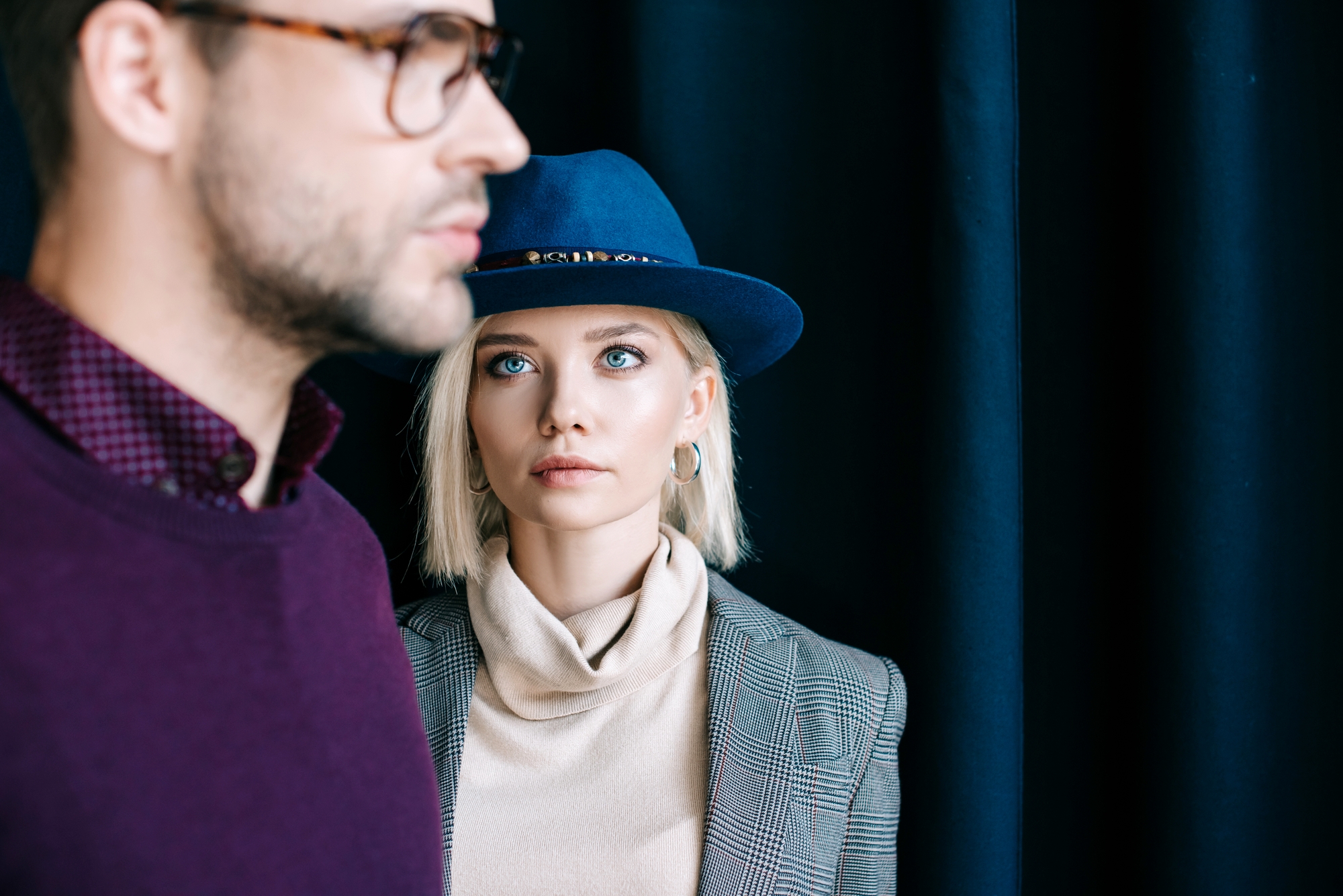 A woman with light hair, wearing a blue hat and a grey checkered blazer over a beige turtleneck, gazes forward with a serious expression. A man wearing glasses and a purple shirt is in the foreground, slightly out of focus, facing away from her.