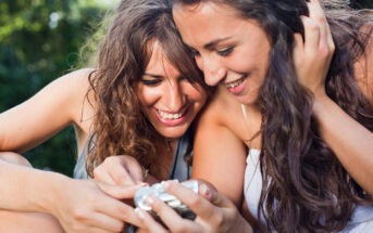 Two women with long, wavy hair are closely looking at a silver camera or a device in one woman's hand. They appear to be outdoors, likely in a park, smiling and enjoying a moment together in the sunlight. Lush greenery is visible in the background.