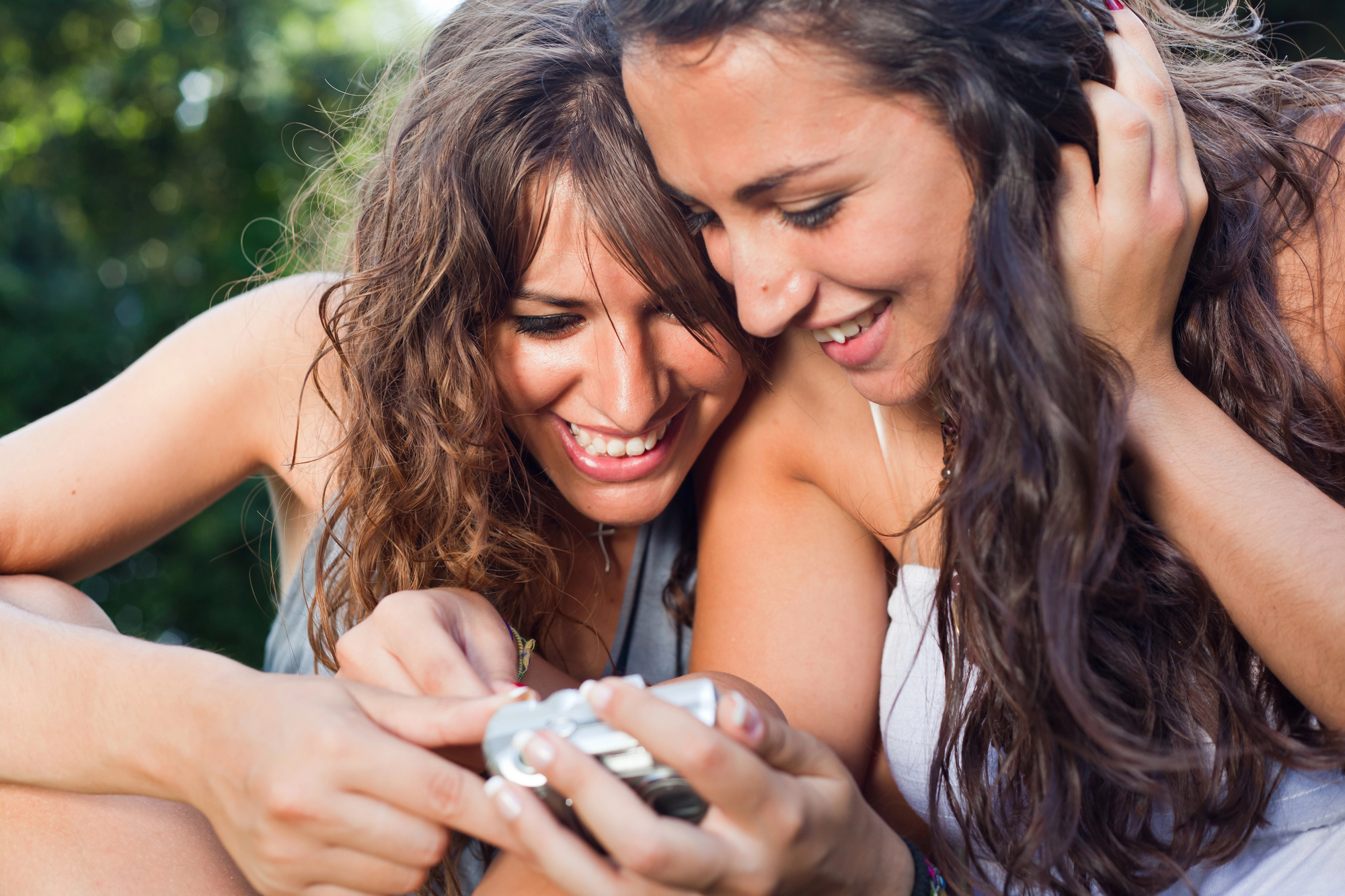 Two women with long, wavy hair are closely looking at a silver camera or a device in one woman's hand. They appear to be outdoors, likely in a park, smiling and enjoying a moment together in the sunlight. Lush greenery is visible in the background.