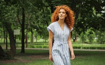 A woman with curly red hair, wearing a striped dress, is walking through a green park. She appears relaxed and content as she strolls along a tree-lined path with lush foliage in the background.