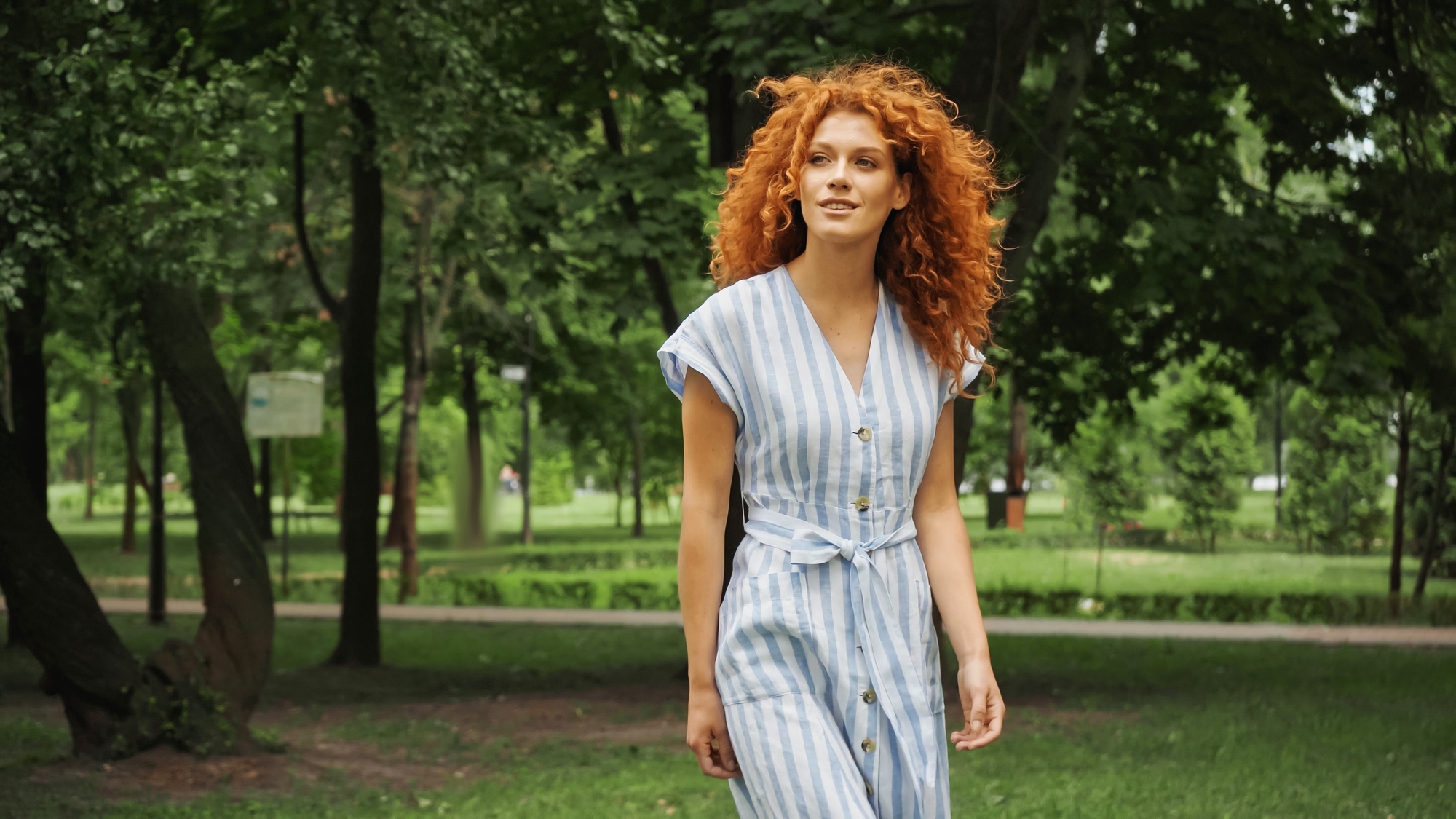 A woman with curly red hair, wearing a striped dress, is walking through a green park. She appears relaxed and content as she strolls along a tree-lined path with lush foliage in the background.