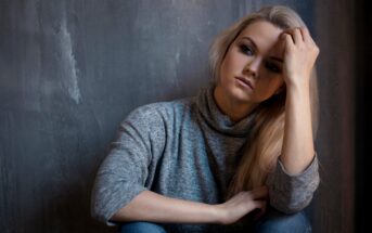 A woman with long blonde hair is sitting against a textured gray wall. She wears a light gray turtleneck sweater and jeans, gazing thoughtfully with one hand resting on her head and the other on her knee. The lighting is soft and subdued.
