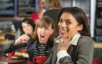 Three women are in a crowded restaurant. The focus is on a smiling woman in a gray blazer in the foreground. Behind her, a woman is angrily shouting and gesturing, while another woman looks on with a concerned expression. Coffee cups and food are on the table.