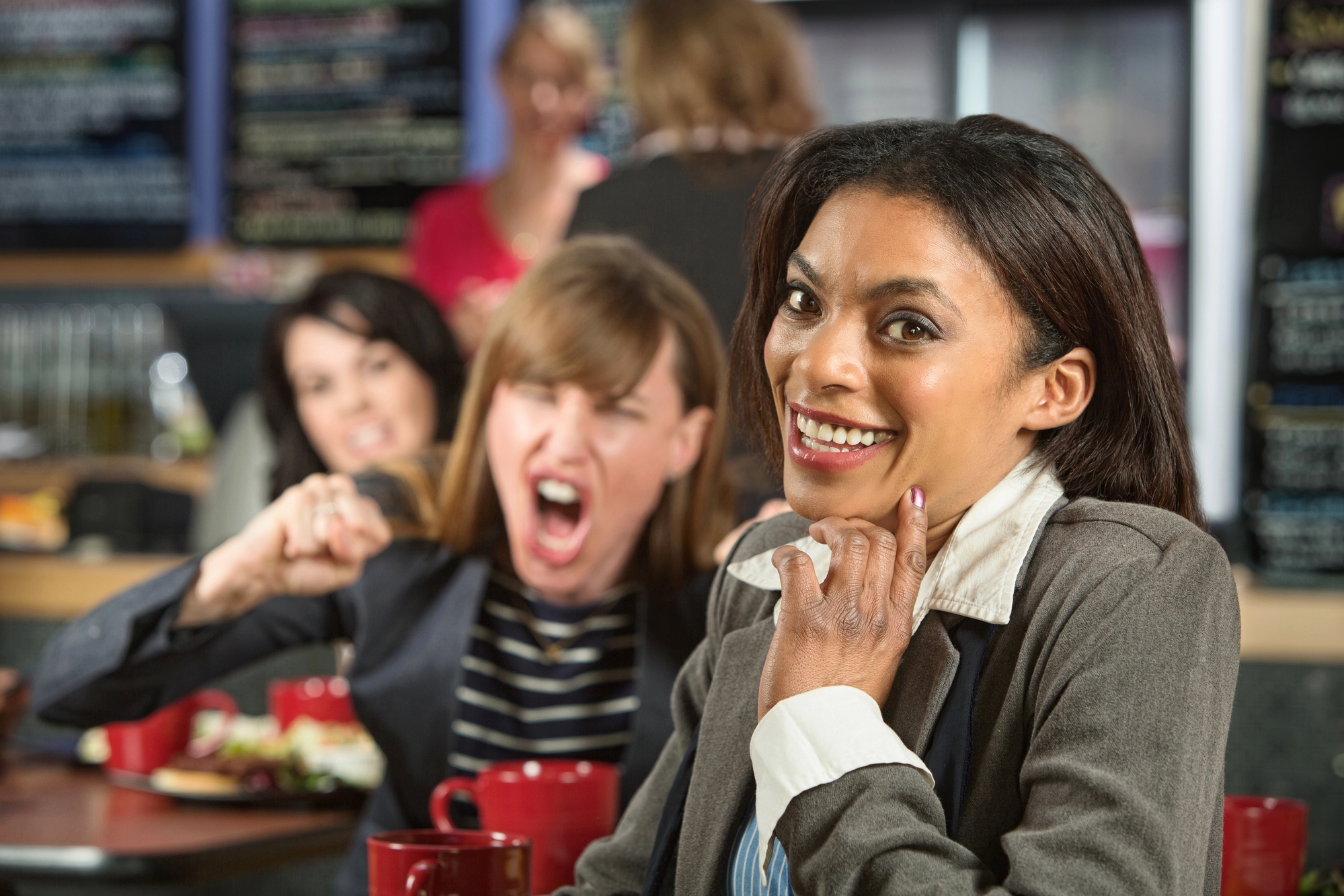 Three women are in a crowded restaurant. The focus is on a smiling woman in a gray blazer in the foreground. Behind her, a woman is angrily shouting and gesturing, while another woman looks on with a concerned expression. Coffee cups and food are on the table.