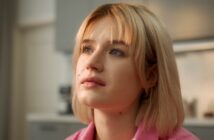 A young woman with short blonde hair gazes thoughtfully upward. She is indoors with a softly lit background, and wears a light pink top.