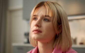 A young woman with short blonde hair gazes thoughtfully upward. She is indoors with a softly lit background, and wears a light pink top.