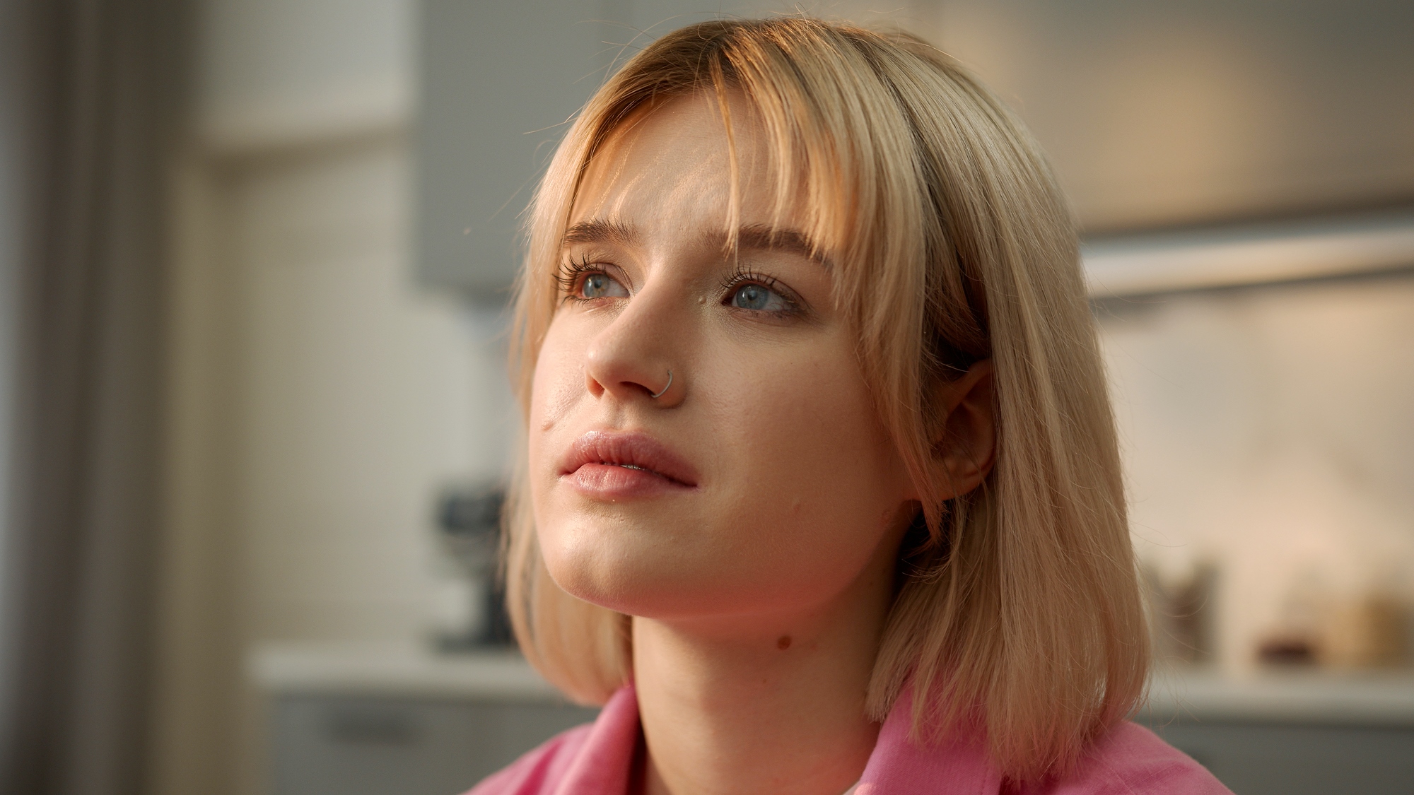 A young woman with short blonde hair gazes thoughtfully upward. She is indoors with a softly lit background, and wears a light pink top.