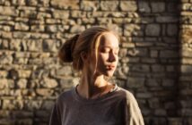 A young woman with blonde hair in a bun stands in front of a sunlit stone wall. She is wearing a grey shirt and has her eyes closed, with the sunlight casting shadows across her face and the wall behind her.