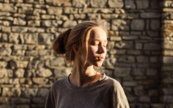 A young woman with blonde hair in a bun stands in front of a sunlit stone wall. She is wearing a grey shirt and has her eyes closed, with the sunlight casting shadows across her face and the wall behind her.