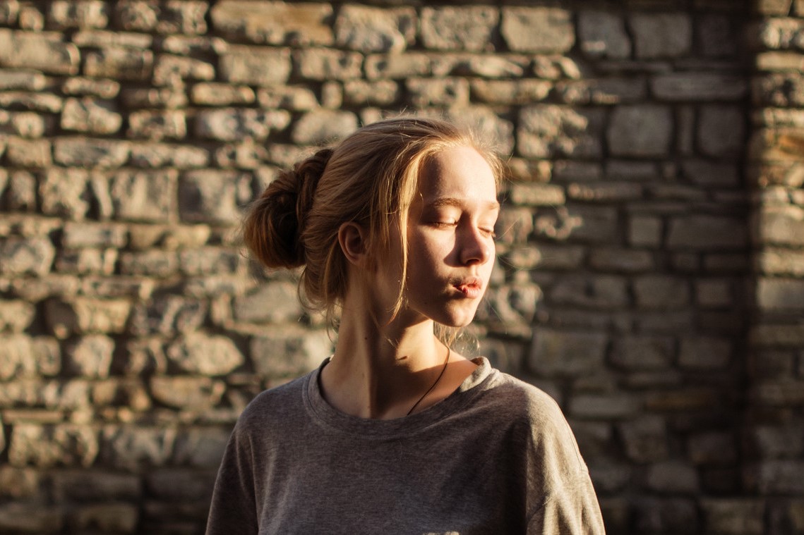 A young woman with blonde hair in a bun stands in front of a sunlit stone wall. She is wearing a grey shirt and has her eyes closed, with the sunlight casting shadows across her face and the wall behind her.