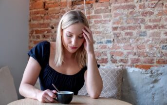 A woman with blonde hair sits at a wooden table in a cafe, holding a black coffee cup with a spoon inside. She rests her head on her hand, looking thoughtful with a pensive expression. The background features an exposed brick wall and a beige cushion.
